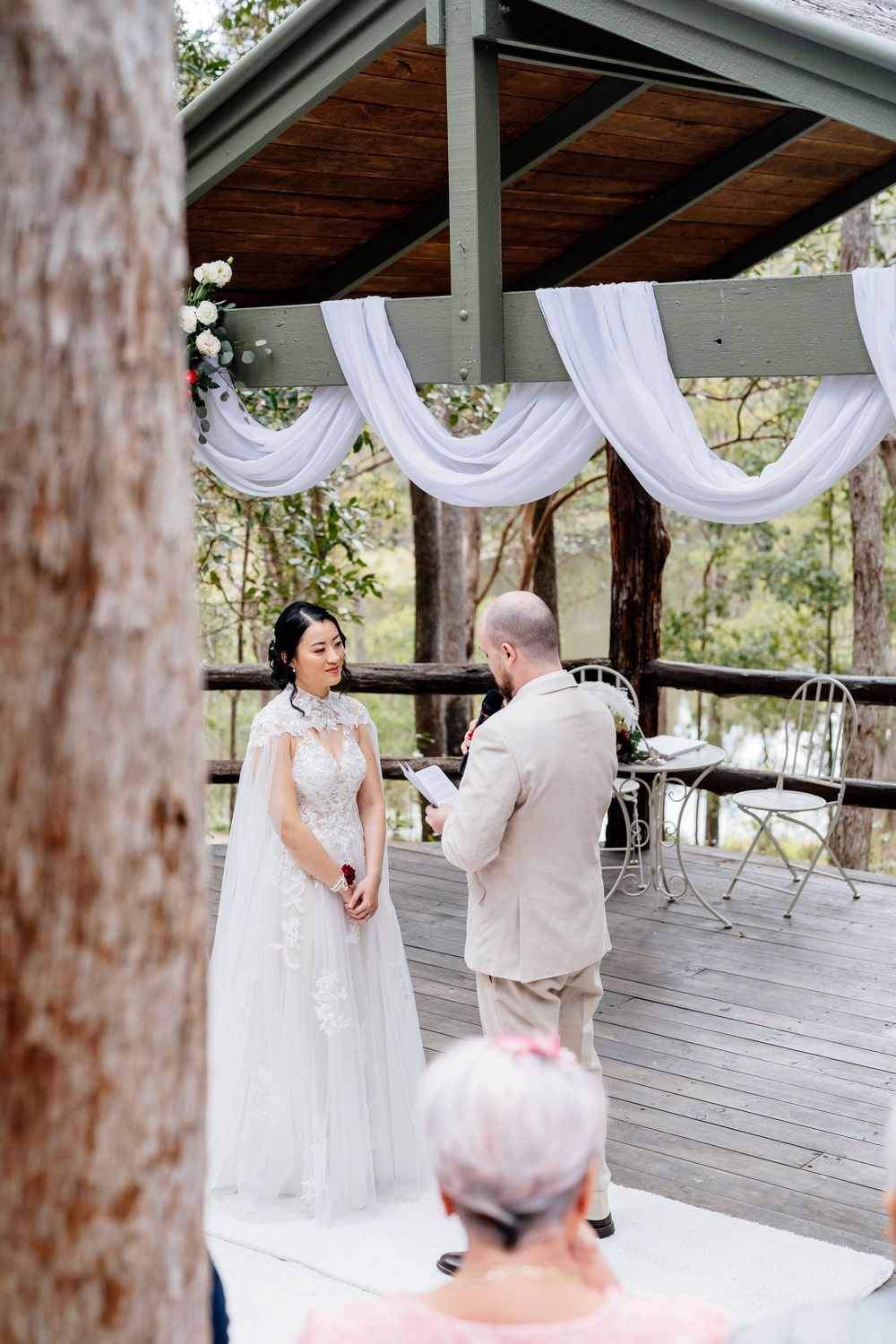 A couple shares a tender moment during their wedding ceremony under a white-draped gazebo in a wooded setting.