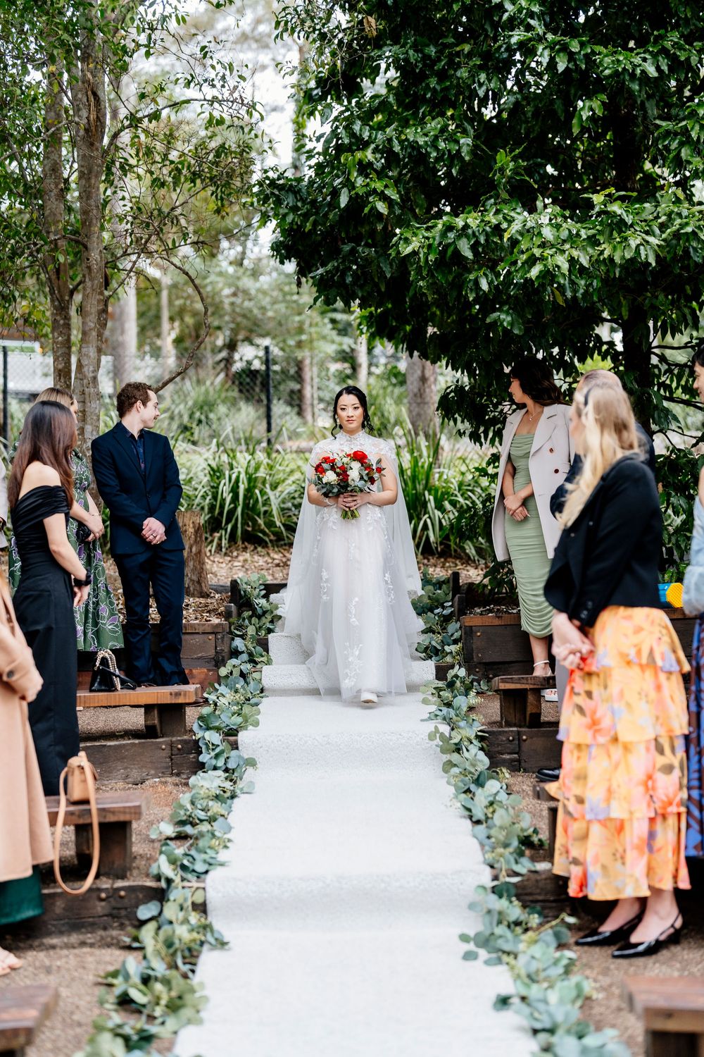 A beautiful outdoor wedding ceremony setup with a white aisle runner decorated with eucalyptus leaves and guests seated on benches.