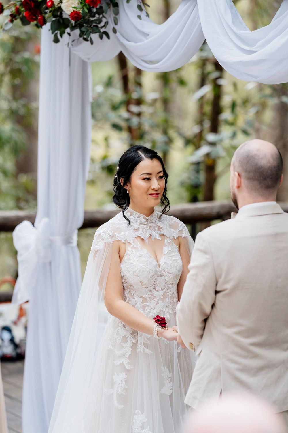 An elegant outdoor wedding ceremony under a white draped arch decorated with red flowers.
