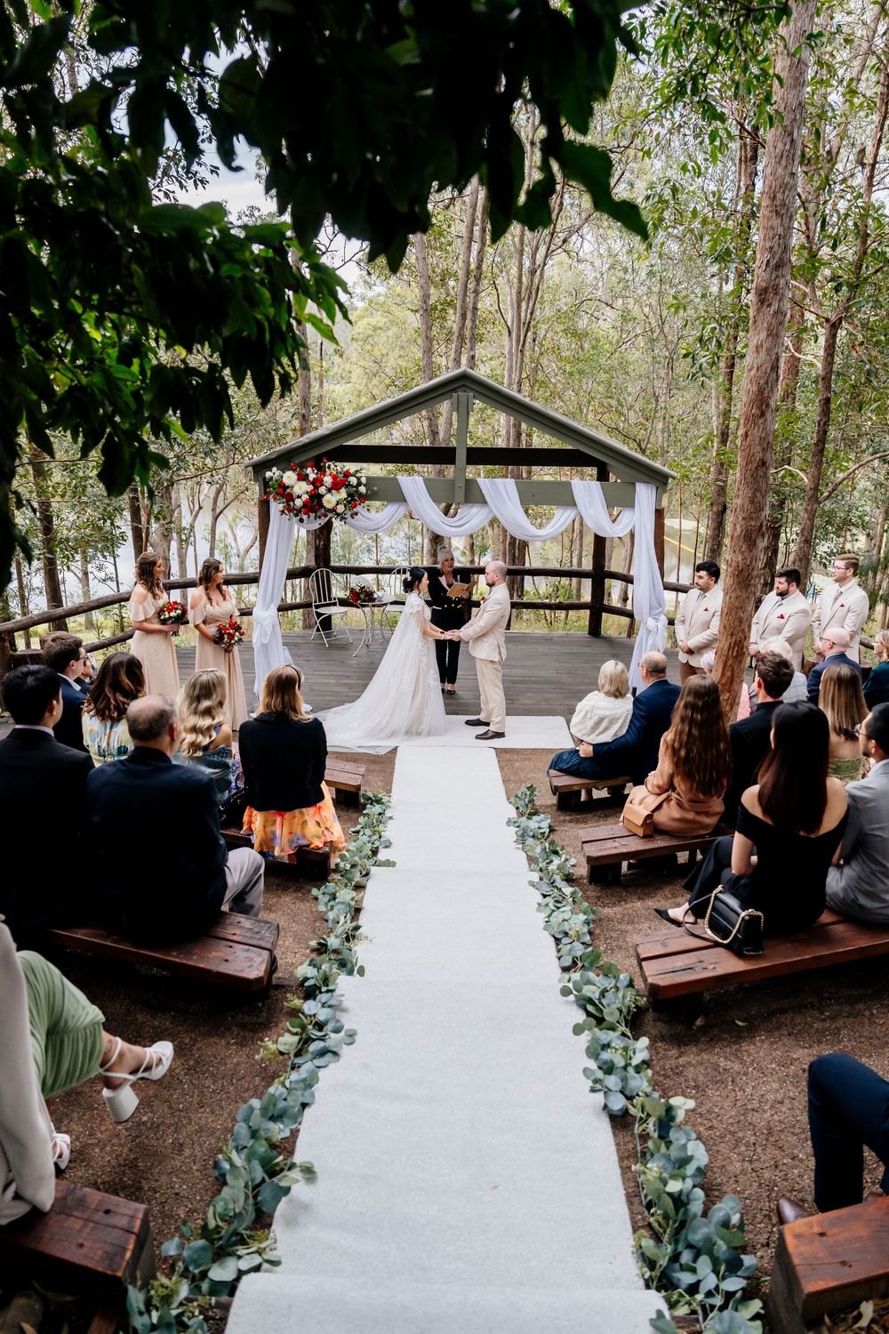 Outdoor forest wedding ceremony with wooden benches and eucalyptus aisle decorations in a rustic woodland setting.