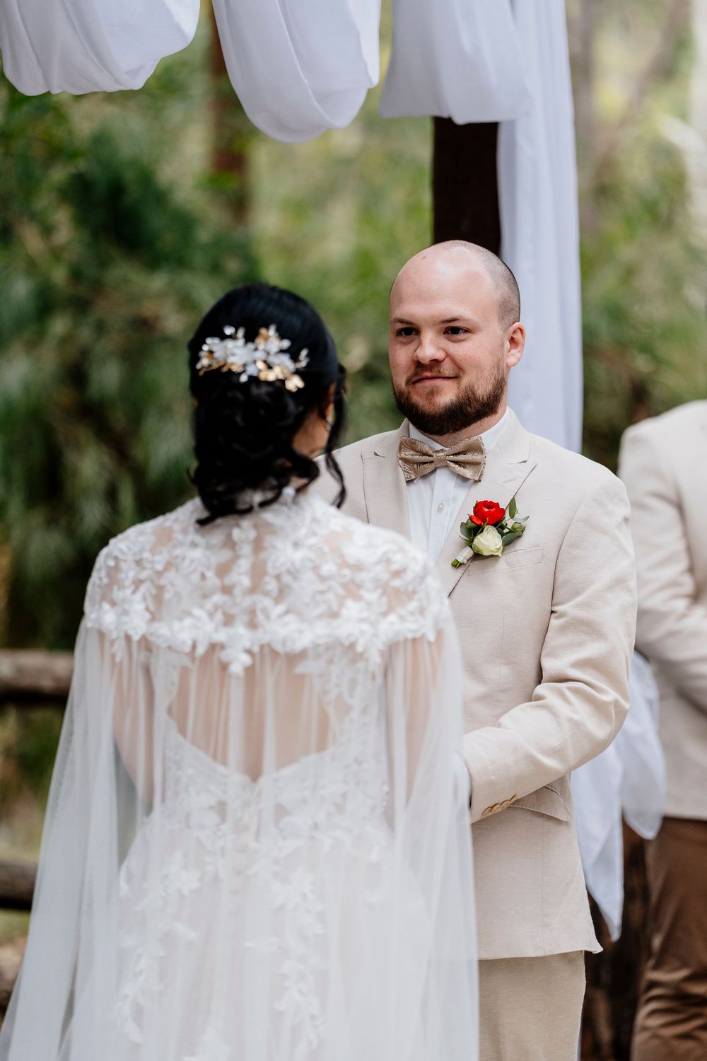 A wedding couple shares an intimate moment during their outdoor ceremony under white fabric draped decorations.
