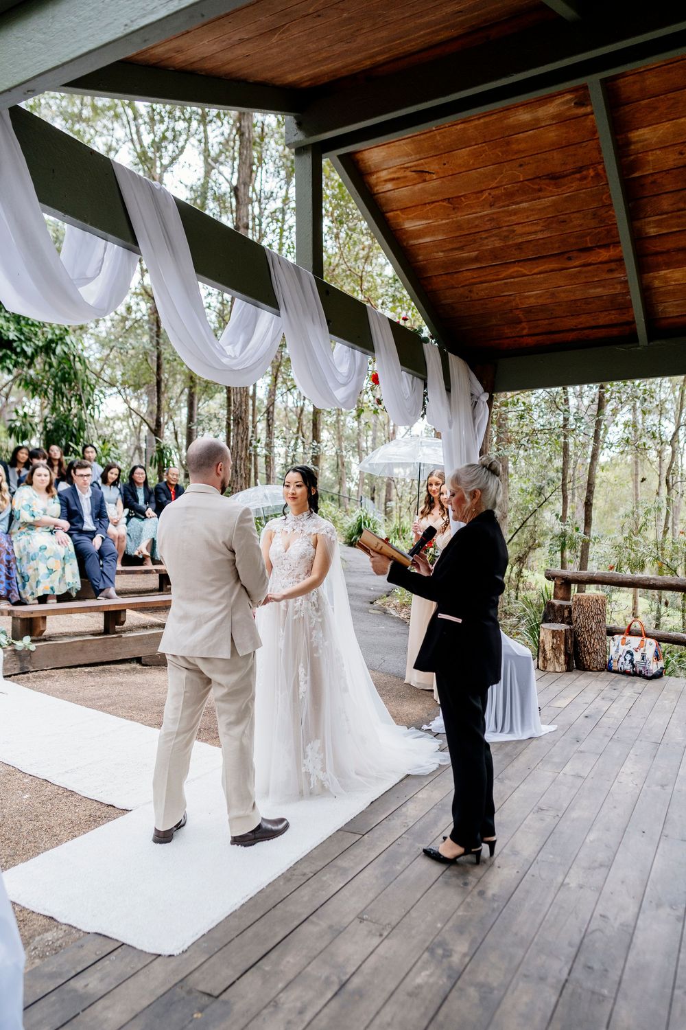 A wedding ceremony takes place on a rustic wooden deck surrounded by forest trees and white fabric draping decorations.
