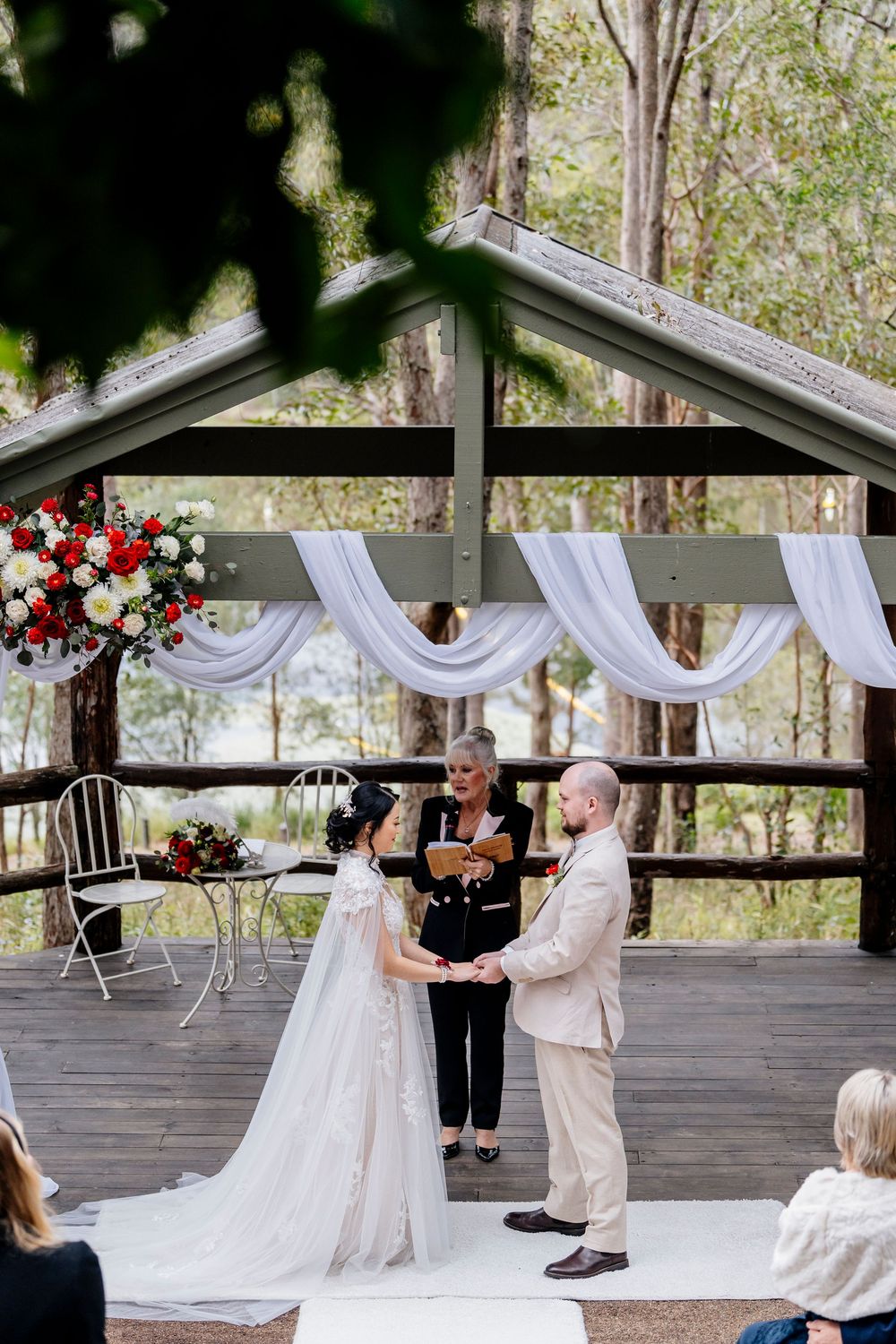 Outdoor wedding ceremony under a rustic wooden pavilion decorated with white draping and red floral arrangements at walkabout creek