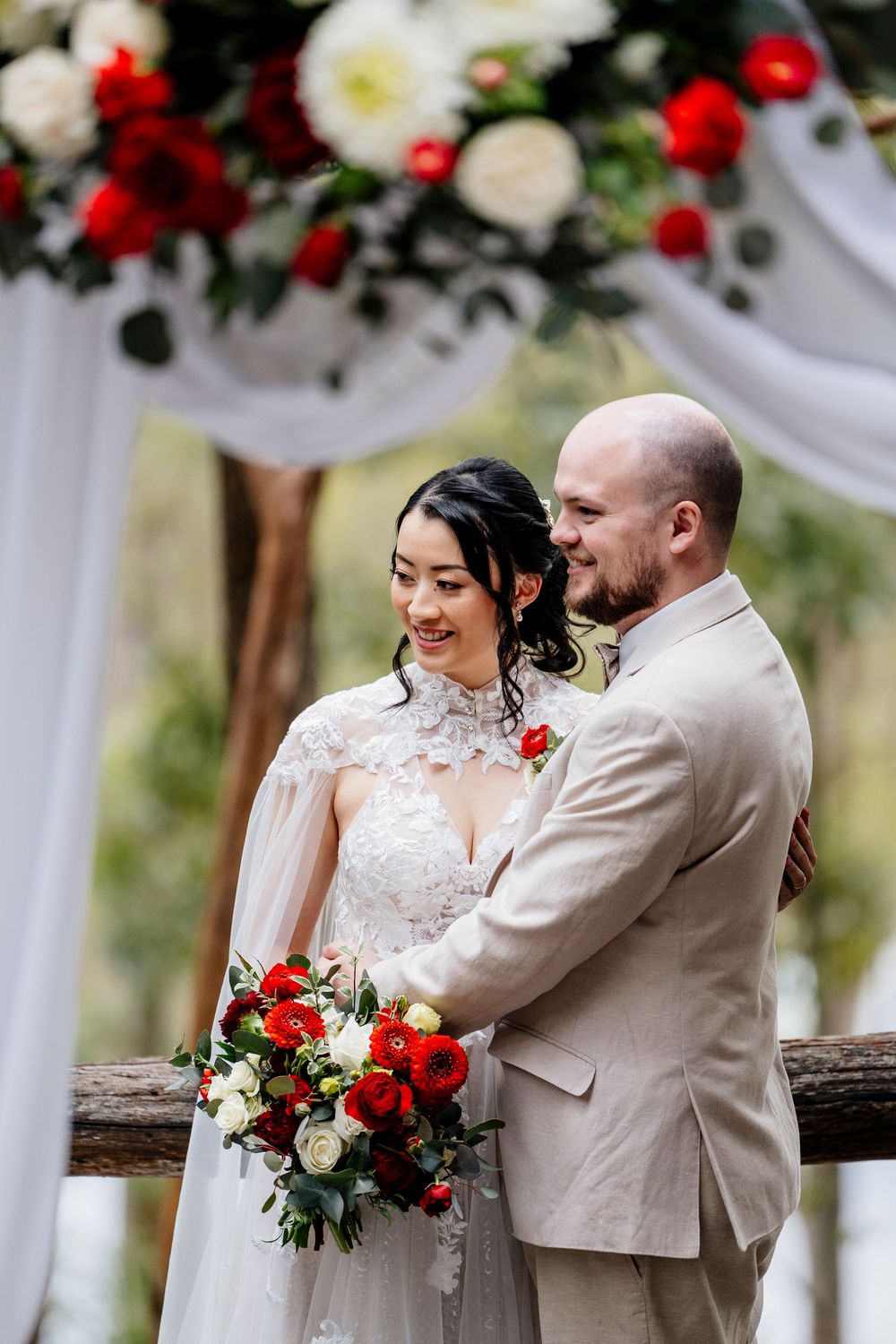 Romantic wedding couple shares joyful moments together against white columns decorated with red and white flowers.