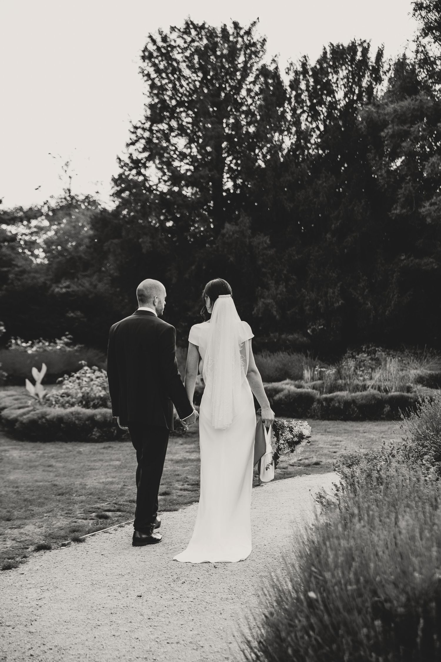 A bride and groom walk together on a path through a natural outdoor setting captured in black and white.