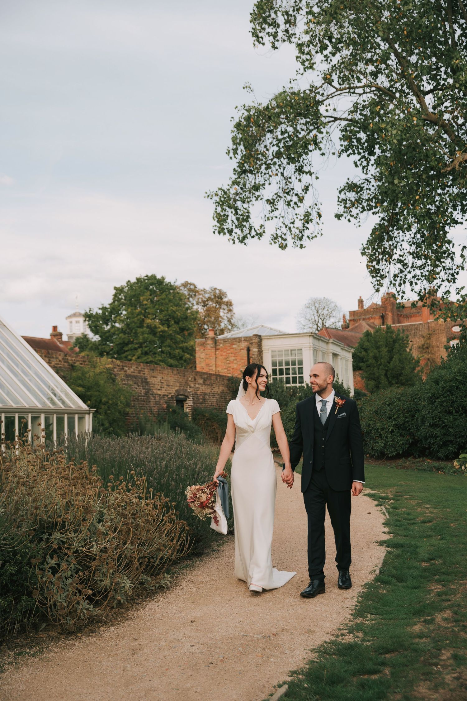 A couple strolls along a garden path with brick buildings and autumn trees in the background.
