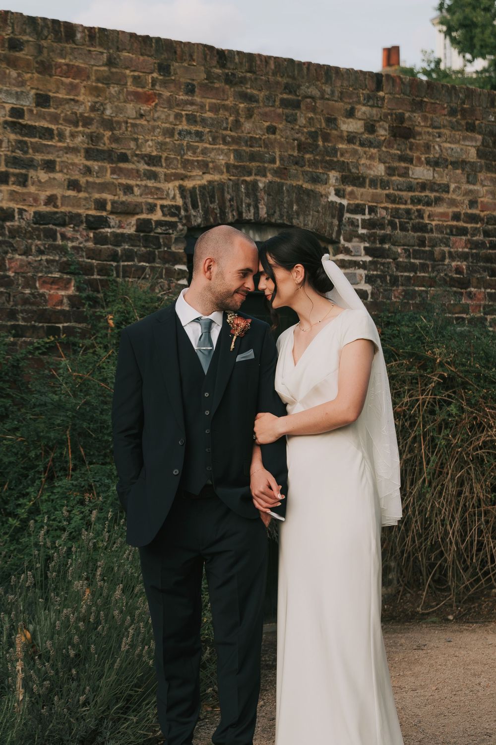 A romantic wedding couple shares an intimate moment against a rustic brick wall in an outdoor garden setting.