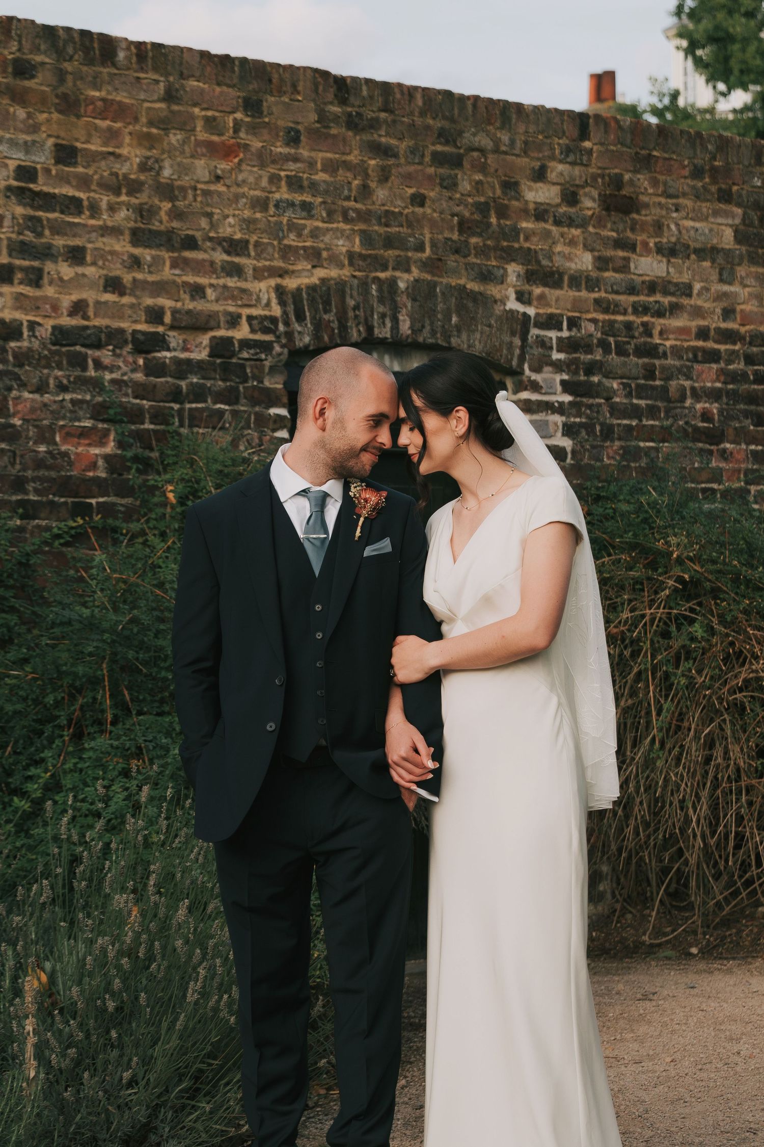 A romantic wedding couple shares an intimate moment against a rustic brick wall in an outdoor garden setting.