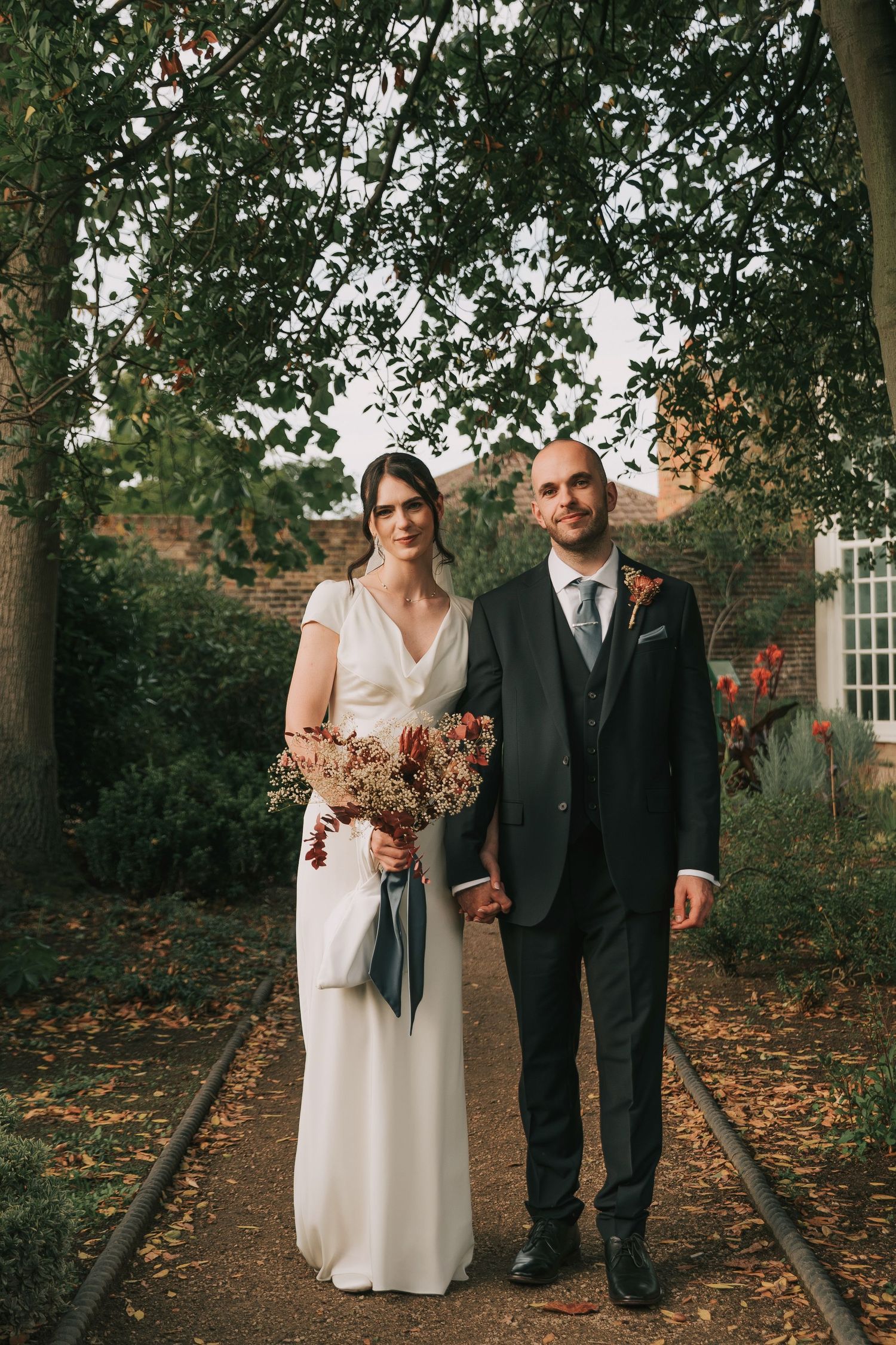 A couple stands together on a tree-lined path covered in autumn leaves during their outdoor fall wedding.