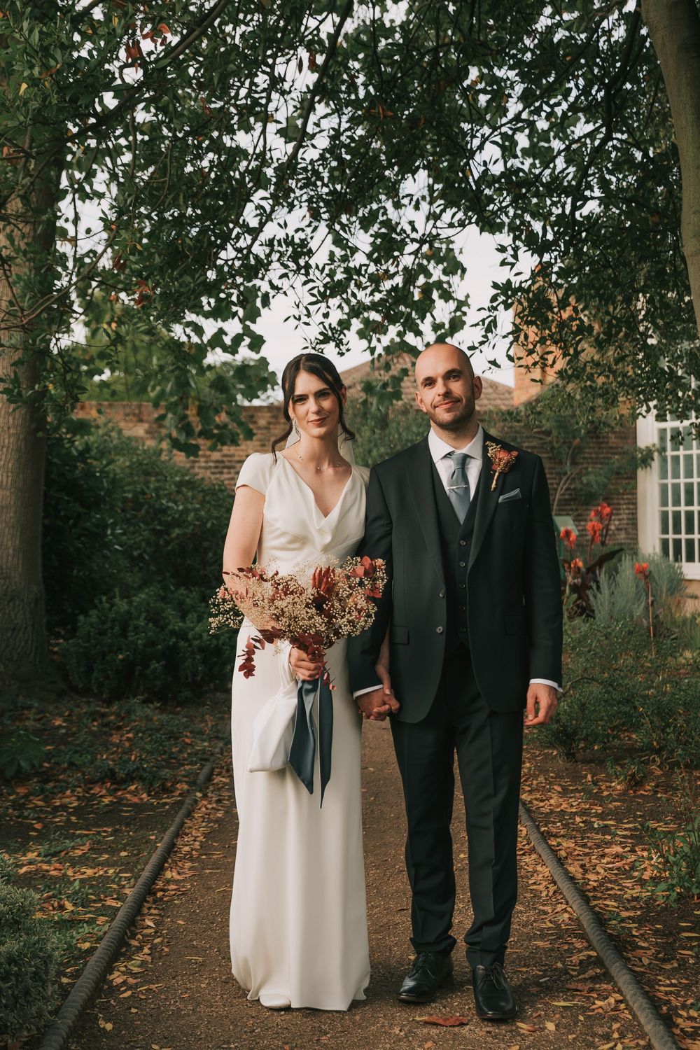 A couple stands together on a tree-lined path covered in autumn leaves during their outdoor fall wedding.