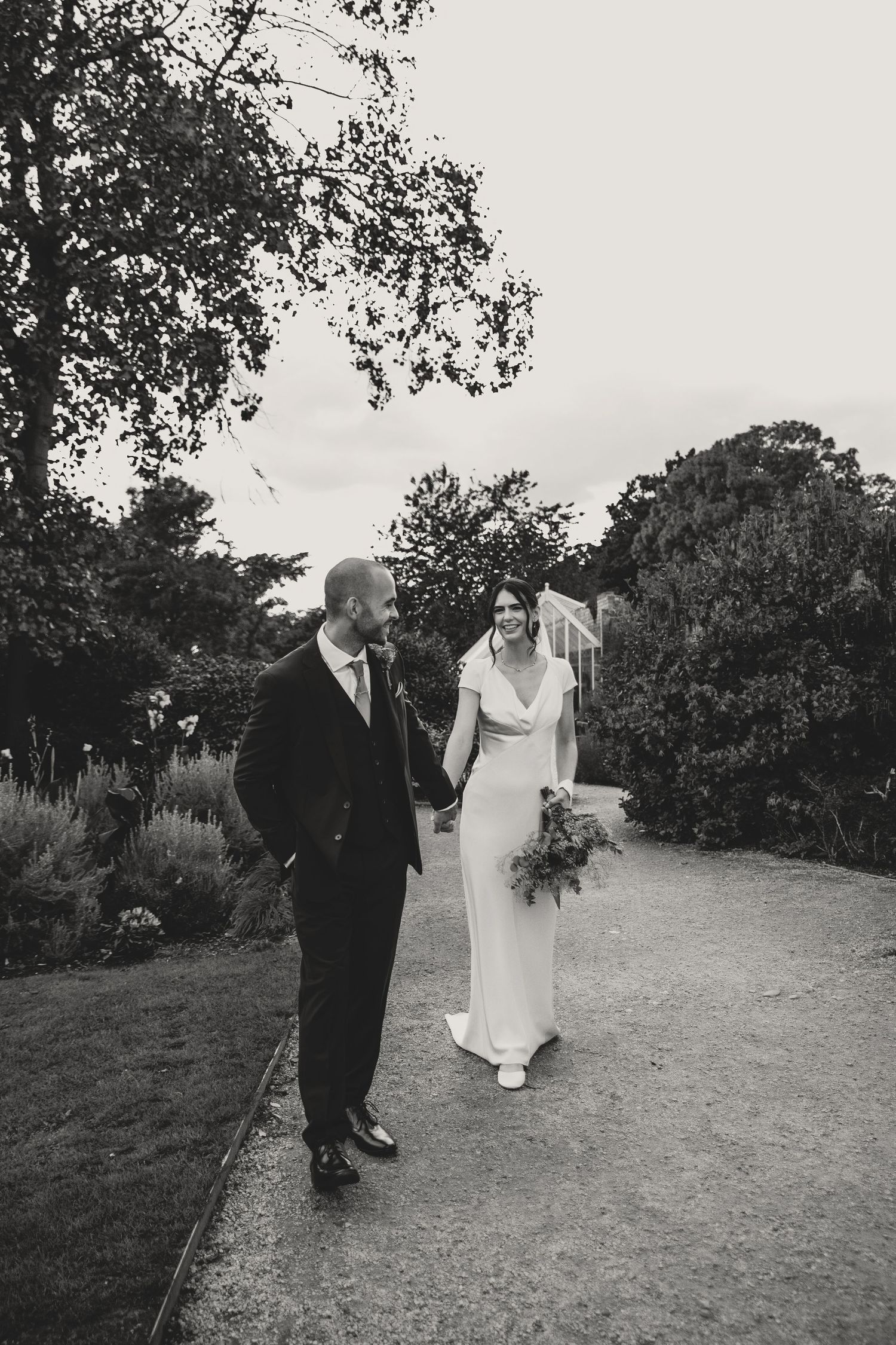 A wedding couple walks together on a winding dirt path surrounded by lush greenery and trees.