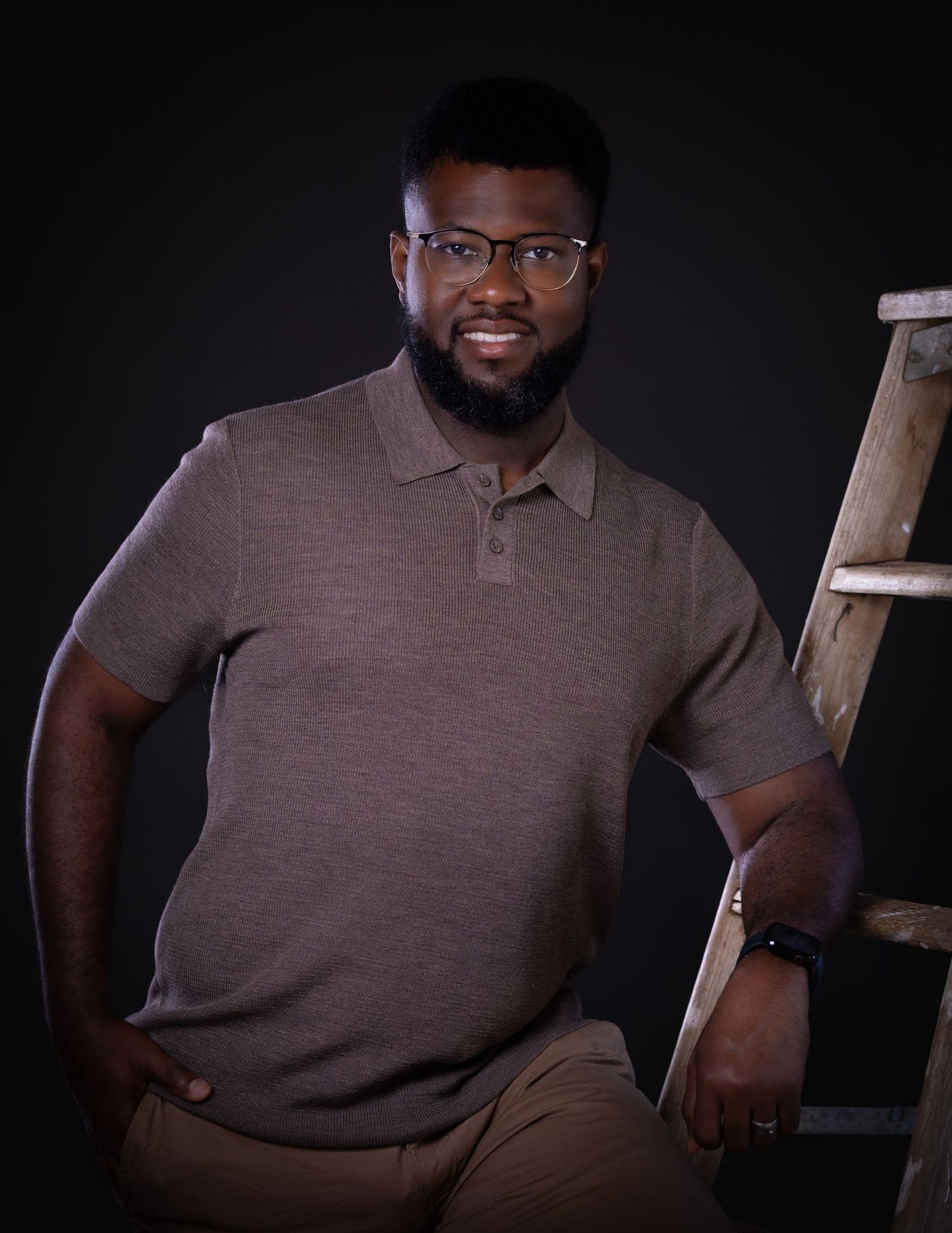 Person in brown polo shirt sitting casually near wooden ladder in moody studio lighting.