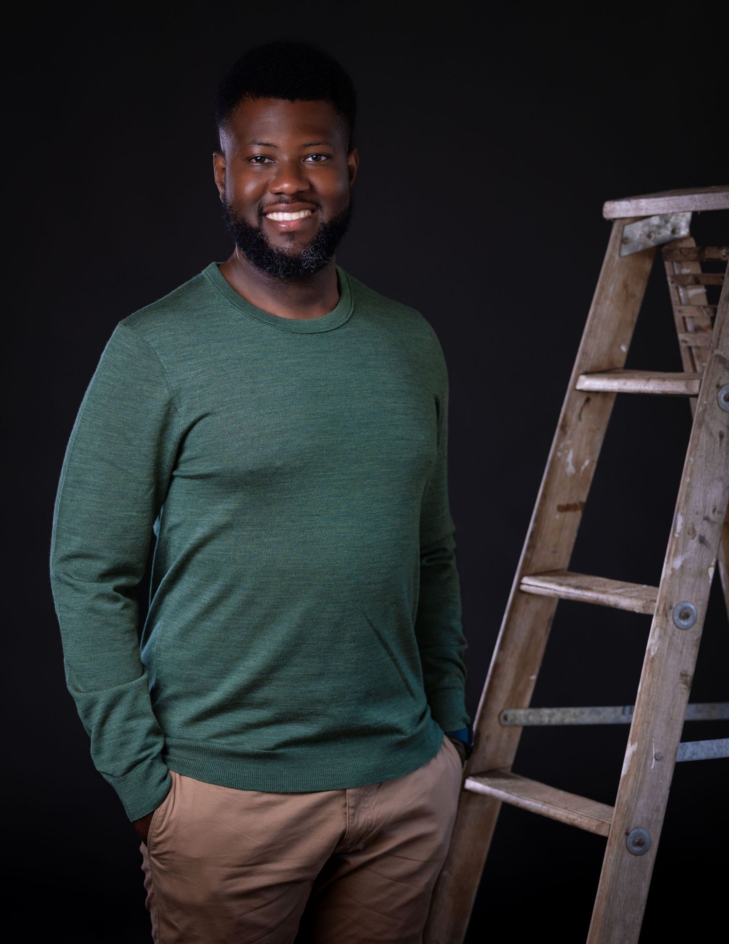 Portrait series against dark background showing casual green sweater and khaki pants with wooden ladder prop.