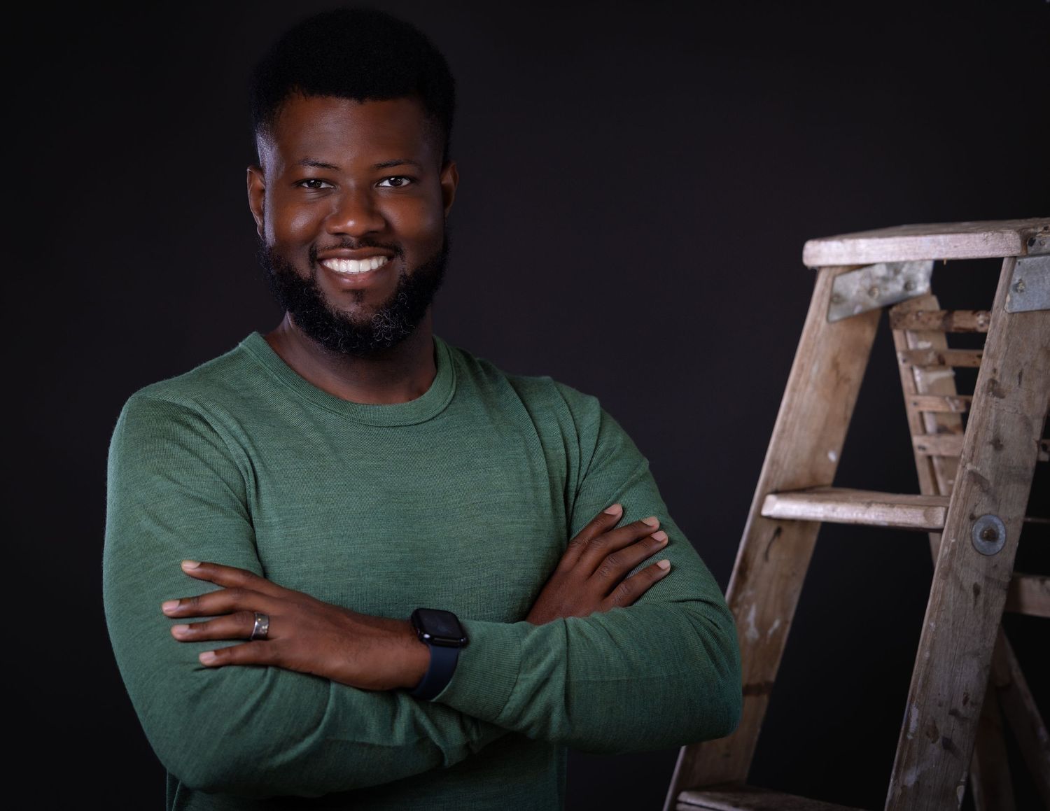 Professional portrait of someone in a green sweater with arms crossed smiling in front of a dark background with a ladder.