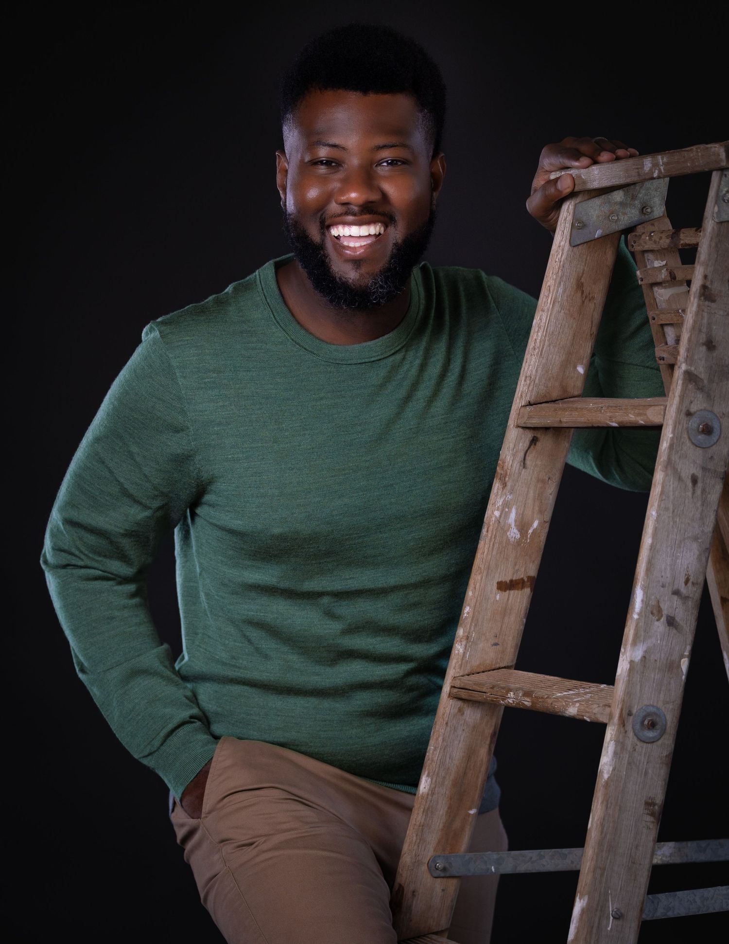 A person in a green sweater and khaki pants sits casually on a wooden ladder against a dark background.