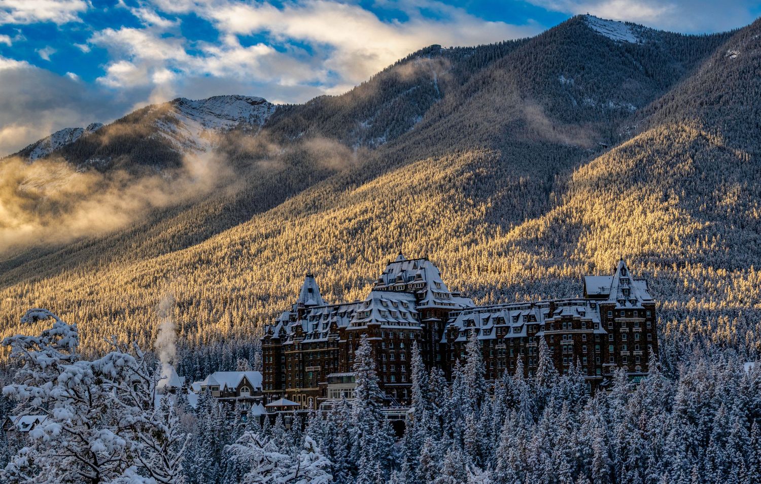 Snow-covered mountain peaks rise above a winter forest landscape under dramatic blue skies and wispy clouds.