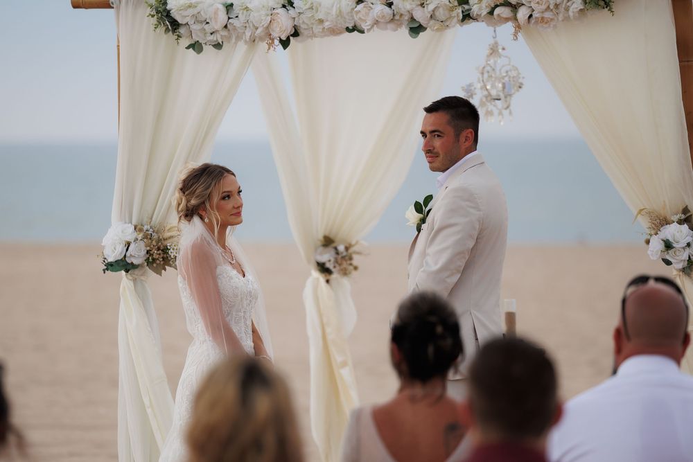 A romantic beach wedding ceremony with white floral arrangements and flowing curtains framing the ocean view.