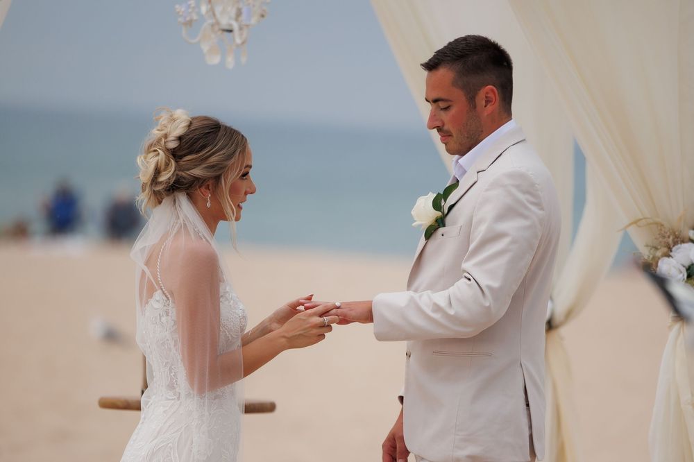 Couple exchanging rings during a beachside wedding ceremony under a white draped canopy.