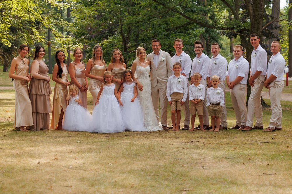 Large wedding party in beige and white attire poses outdoors on grassy lawn.