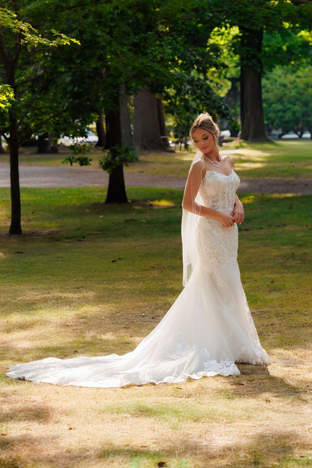 A bride in a flowing white lace wedding dress stands in a sunlit park setting surrounded by trees.