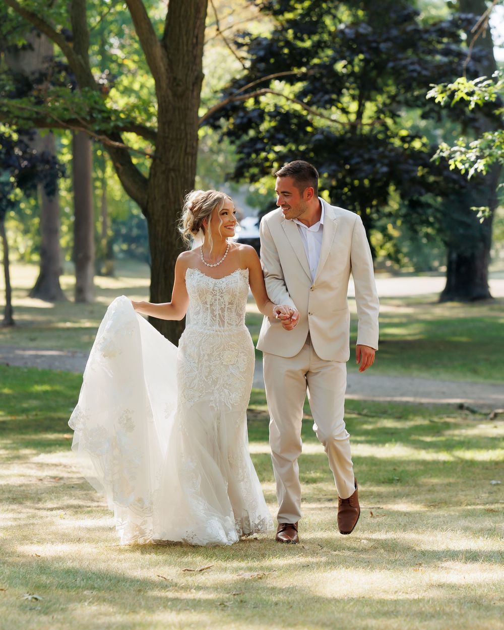 Newlyweds walking together through a sunlit park with trees casting dappled shadows.