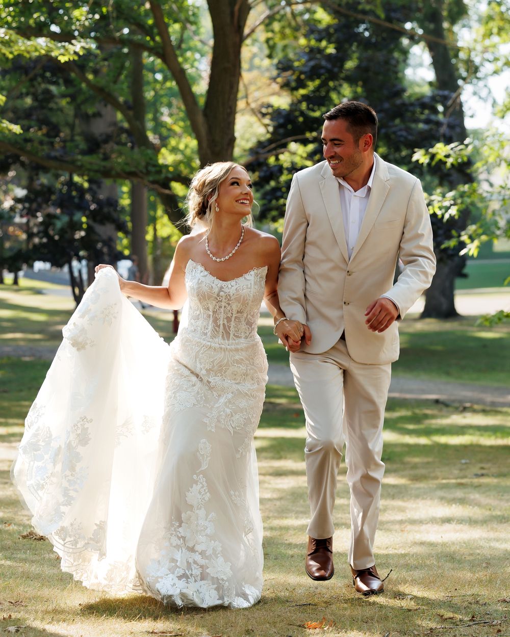 A couple in wedding attire walks together and laughs joyfully in a sunlit outdoor garden setting.