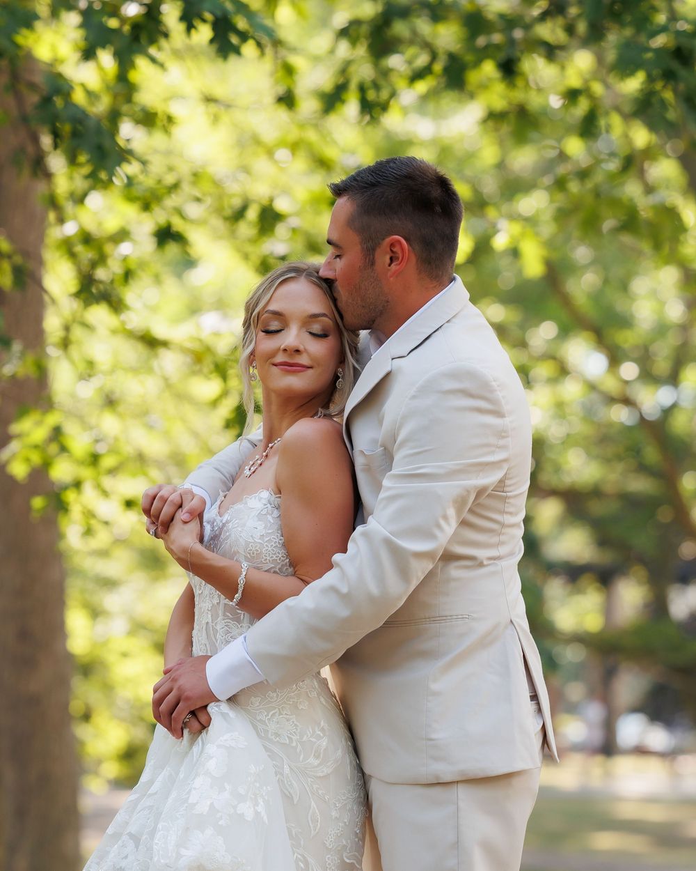 Couple in white wedding attire sharing an intimate moment in a sun-dappled garden setting.