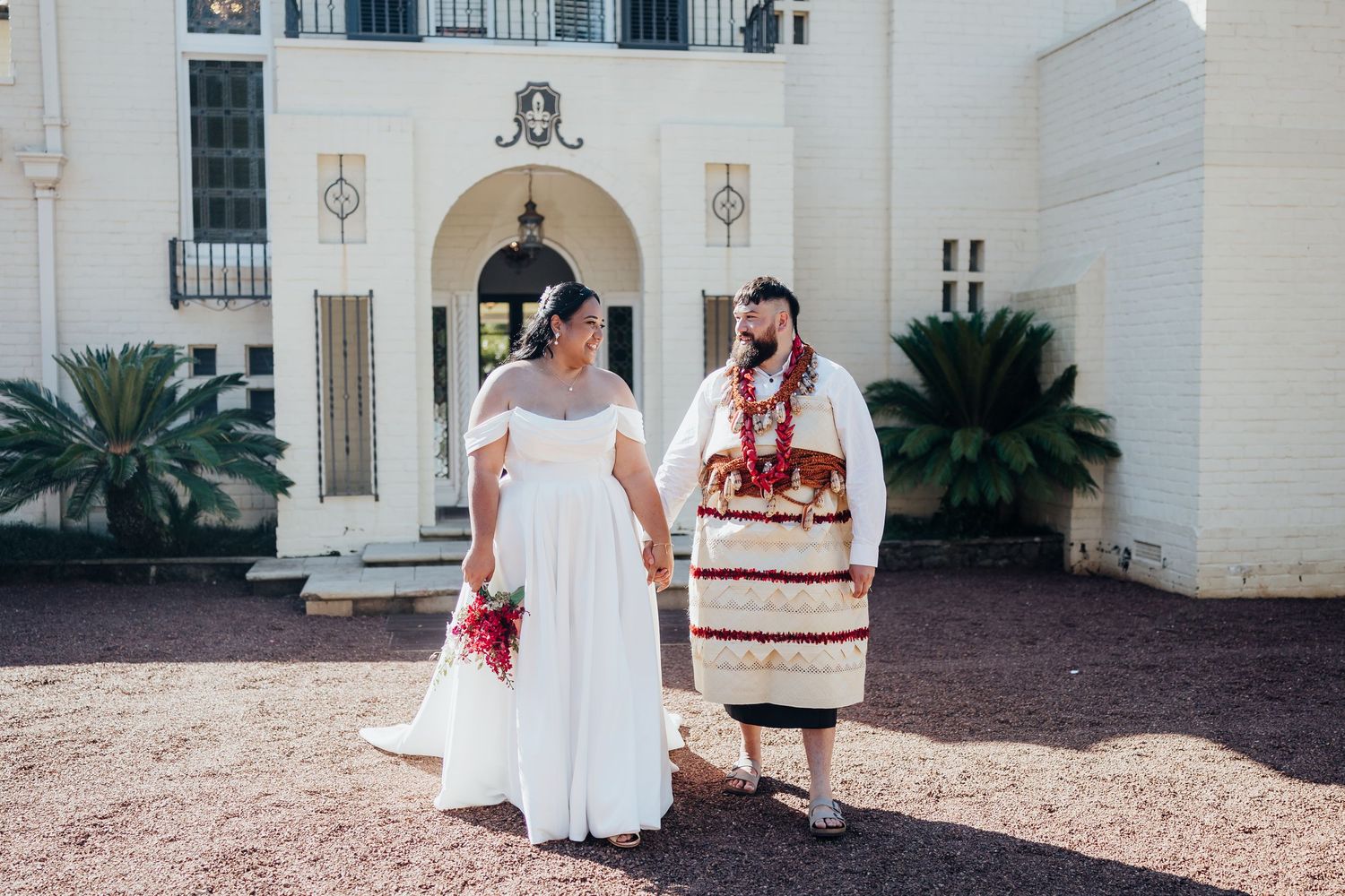 Wedding couple stands on white bridge surrounded by palm trees and lush green landscape.