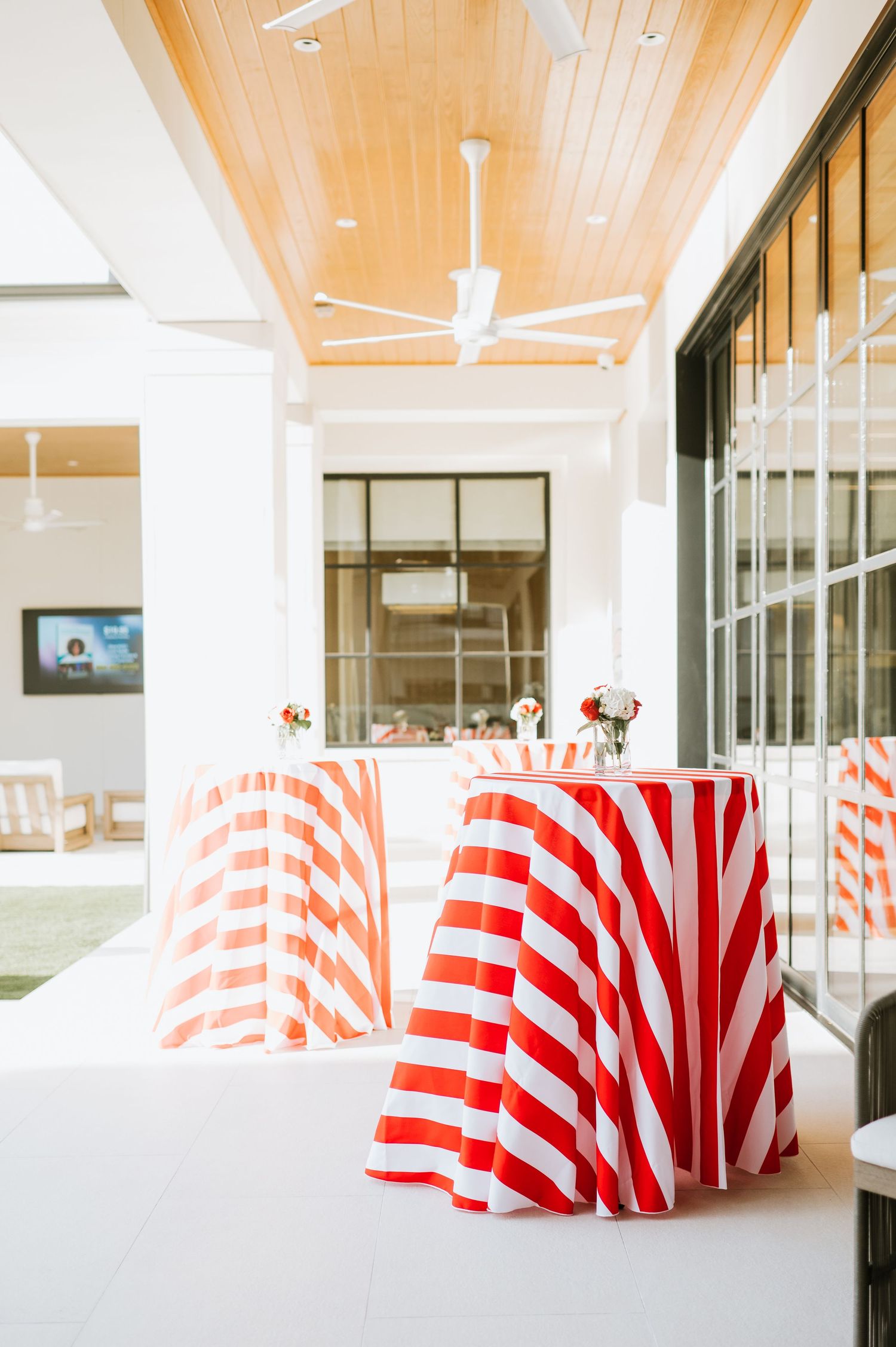 Red and white striped tablecloths on round tables in a modern event space with wooden ceiling panels and glass walls.