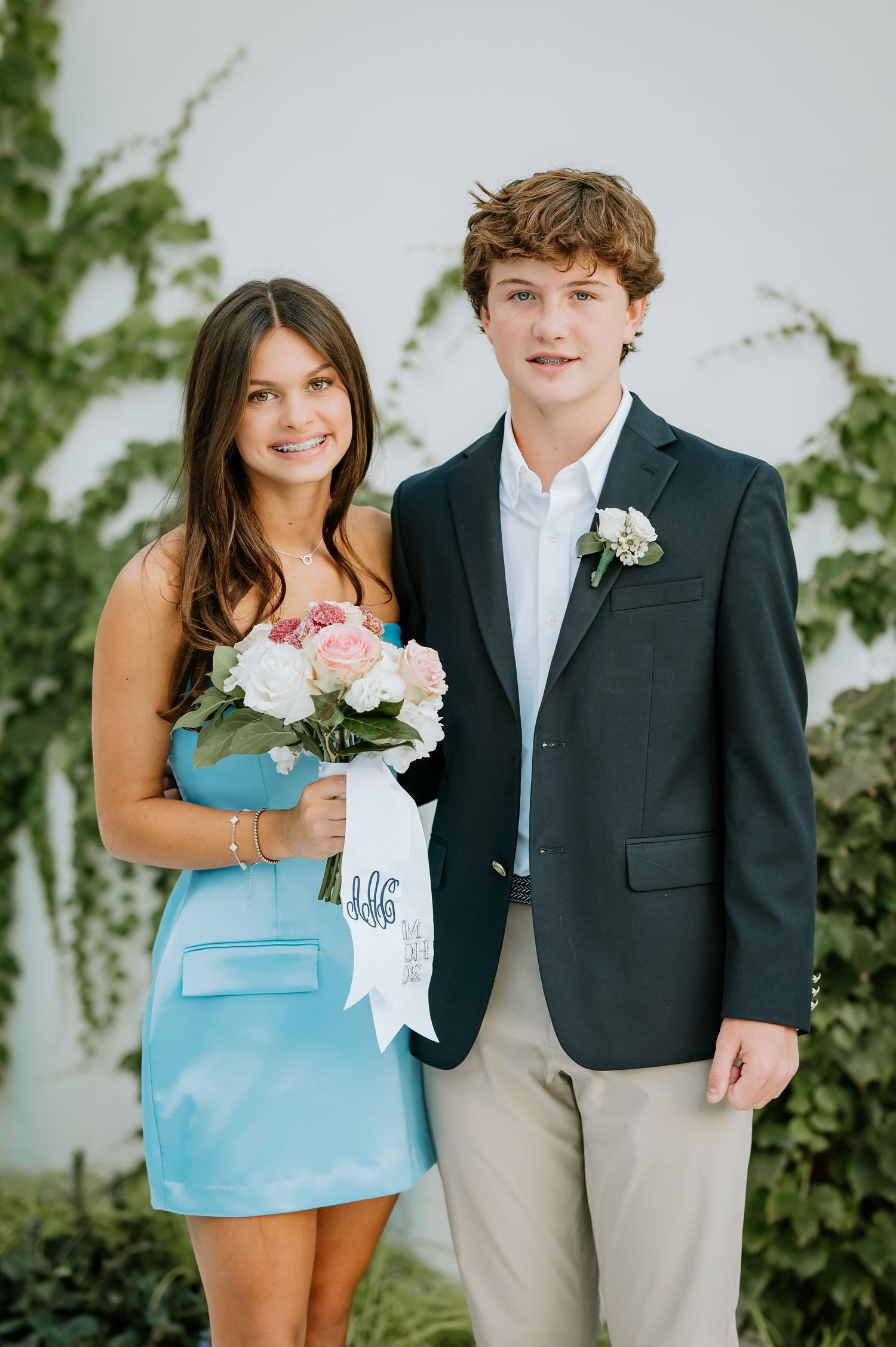 Young couple in formal attire pose together against white wall with green vines for special occasion photos.