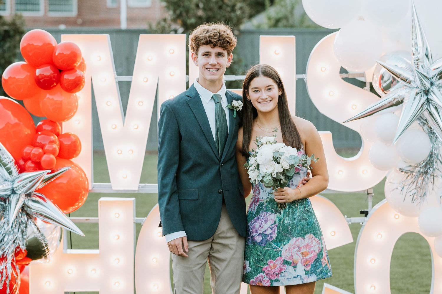 Wedding party poses outdoors with large white letter decor and vibrant red balloon arrangements in the background.