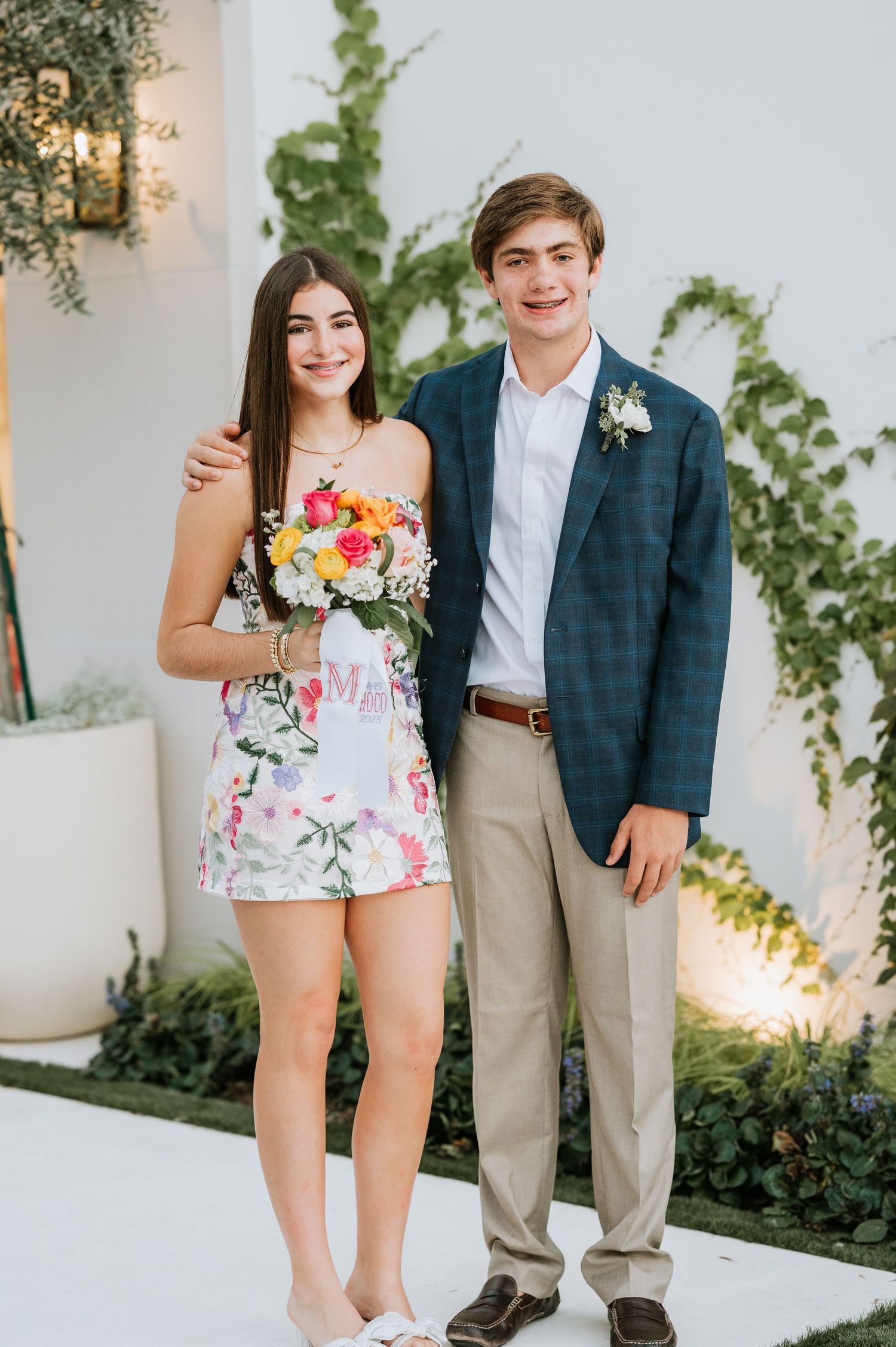 A couple dressed in formal attire pose together at a spring event with floral decorations and greenery.