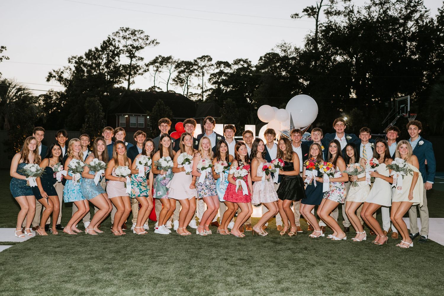 Large group of friends pose together outdoors holding white balloons at dusk.