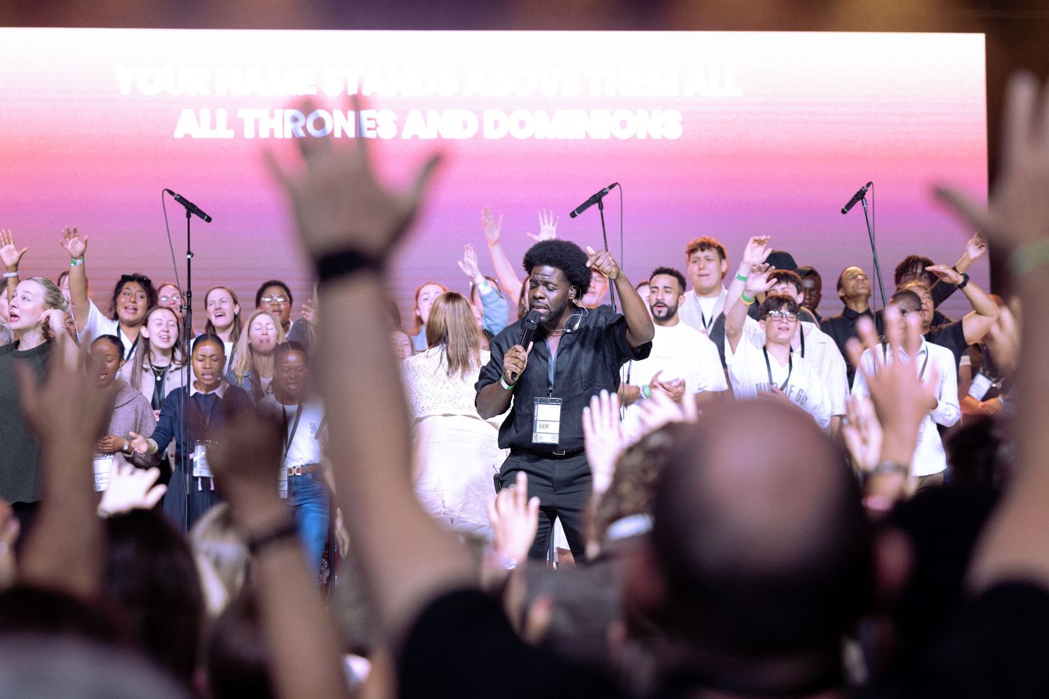 Crowd of people on stage at a large event with audience members raising their hands in foreground.