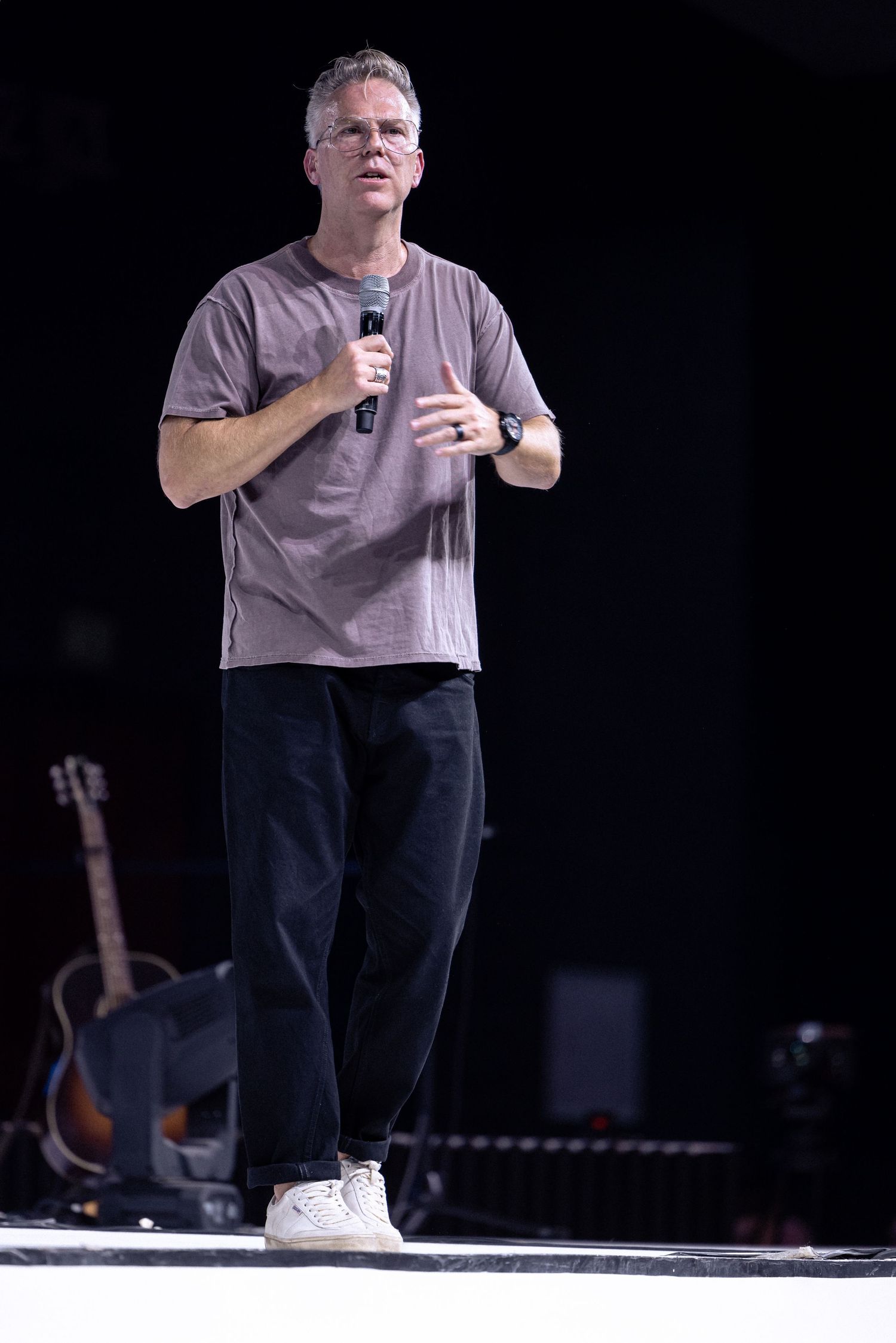 Speaker on stage wearing casual grey t-shirt and dark pants with acoustic guitar visible in background.