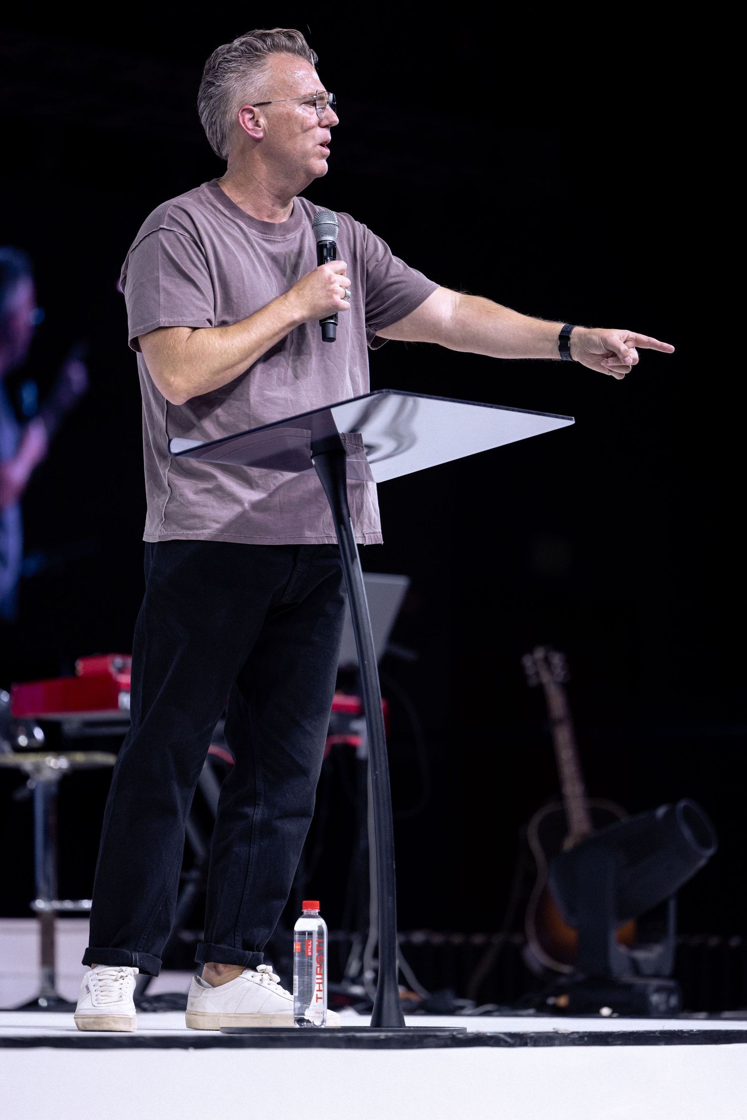 A comedian in a gray t-shirt and black pants performs on stage with animated gestures during a stand-up routine.