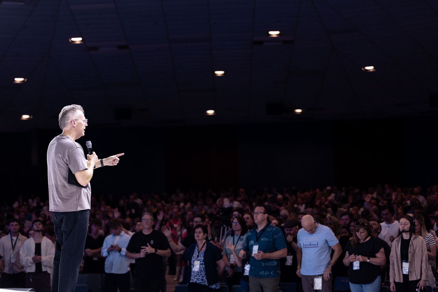 A performer stands on stage addressing a large crowd in a dimly lit auditorium during a live event.