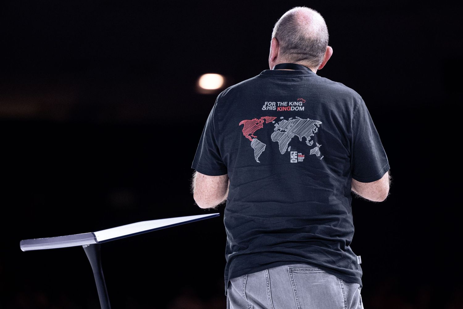 Back view of person wearing black t-shirt with world map design speaking on stage.