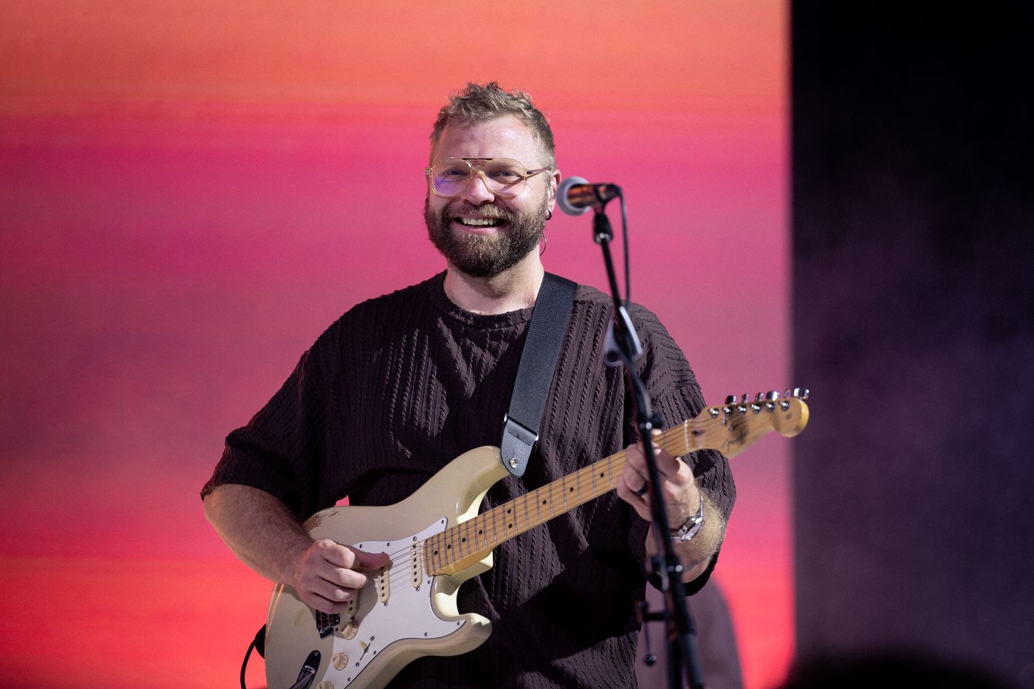 Musicians perform on stage with guitars against a vibrant pink and purple backdrop during a live concert performance.