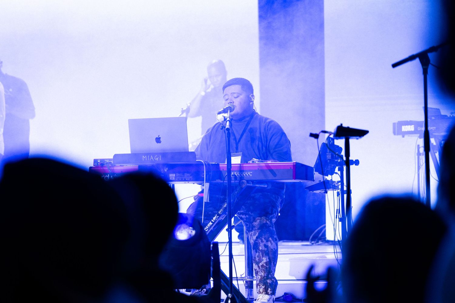 Performer on stage behind keyboard and laptop with blue lighting while silhouetted audience watches from below.
