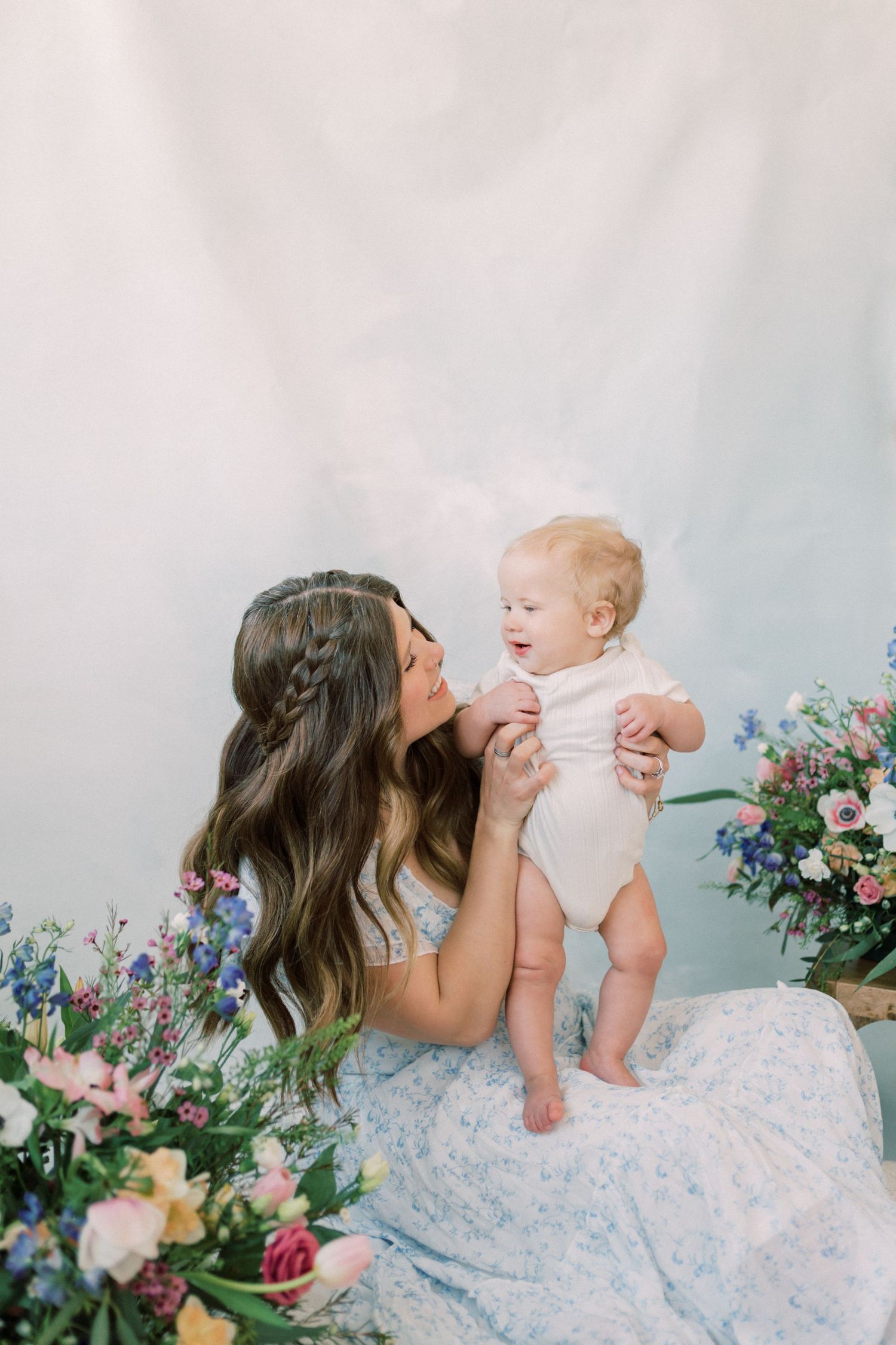 Tender family portrait session with mother and children in soft white and pink dresses against light backdrop with floral decor.