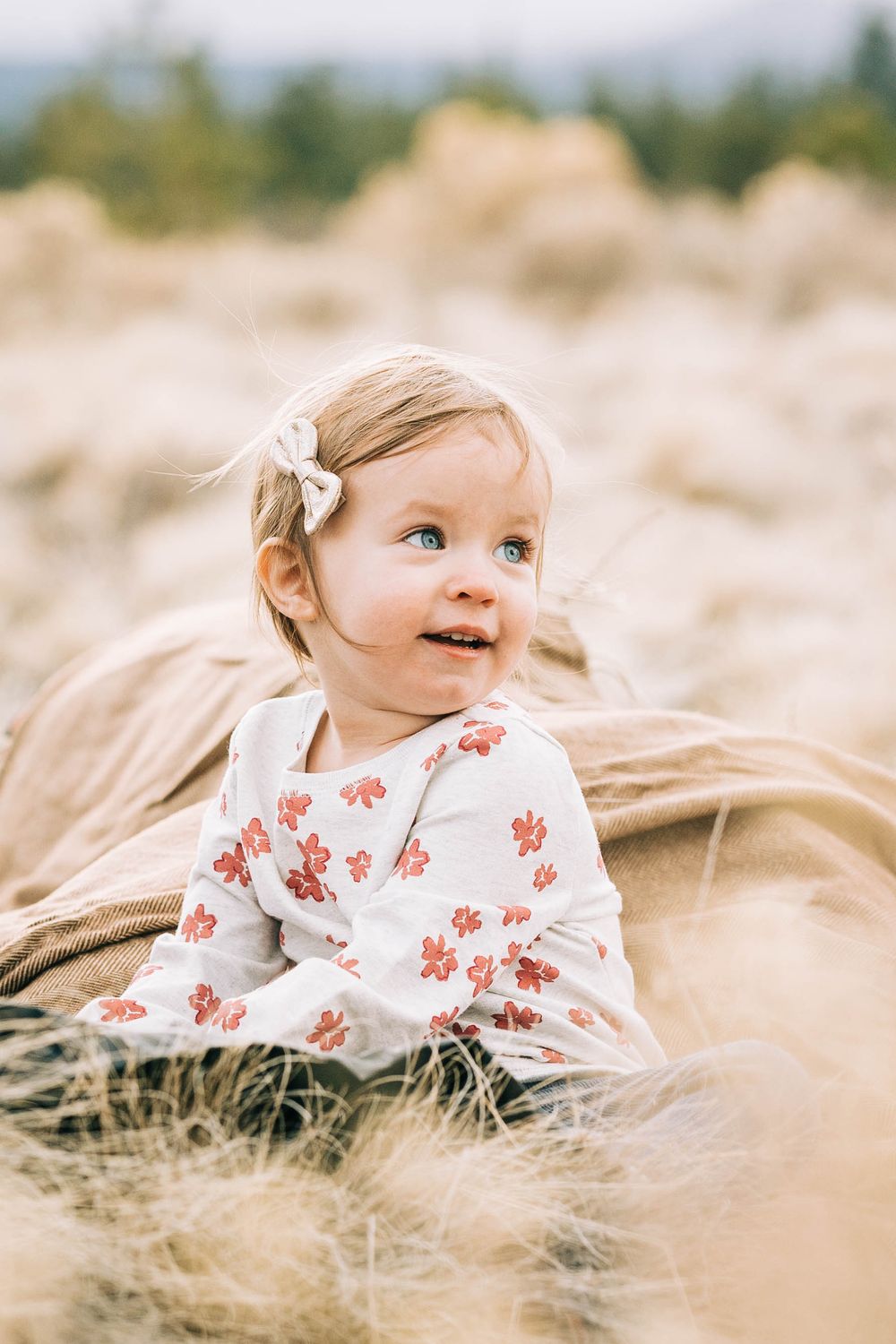 Cute baby girl with blue eyes in floral onesie sitting in golden grass field outdoors.