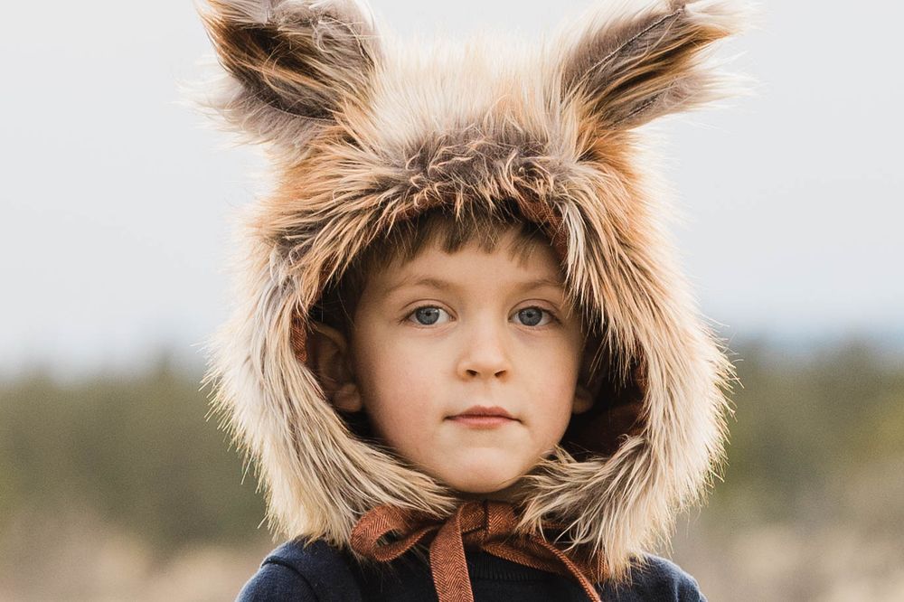 Young child wearing fluffy fox ear hat looking at camera with soft outdoor background.