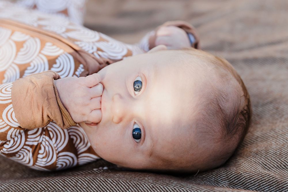 Adorable blue-eyed infant in rainbow patterned sleepsuit lying on grey blanket.