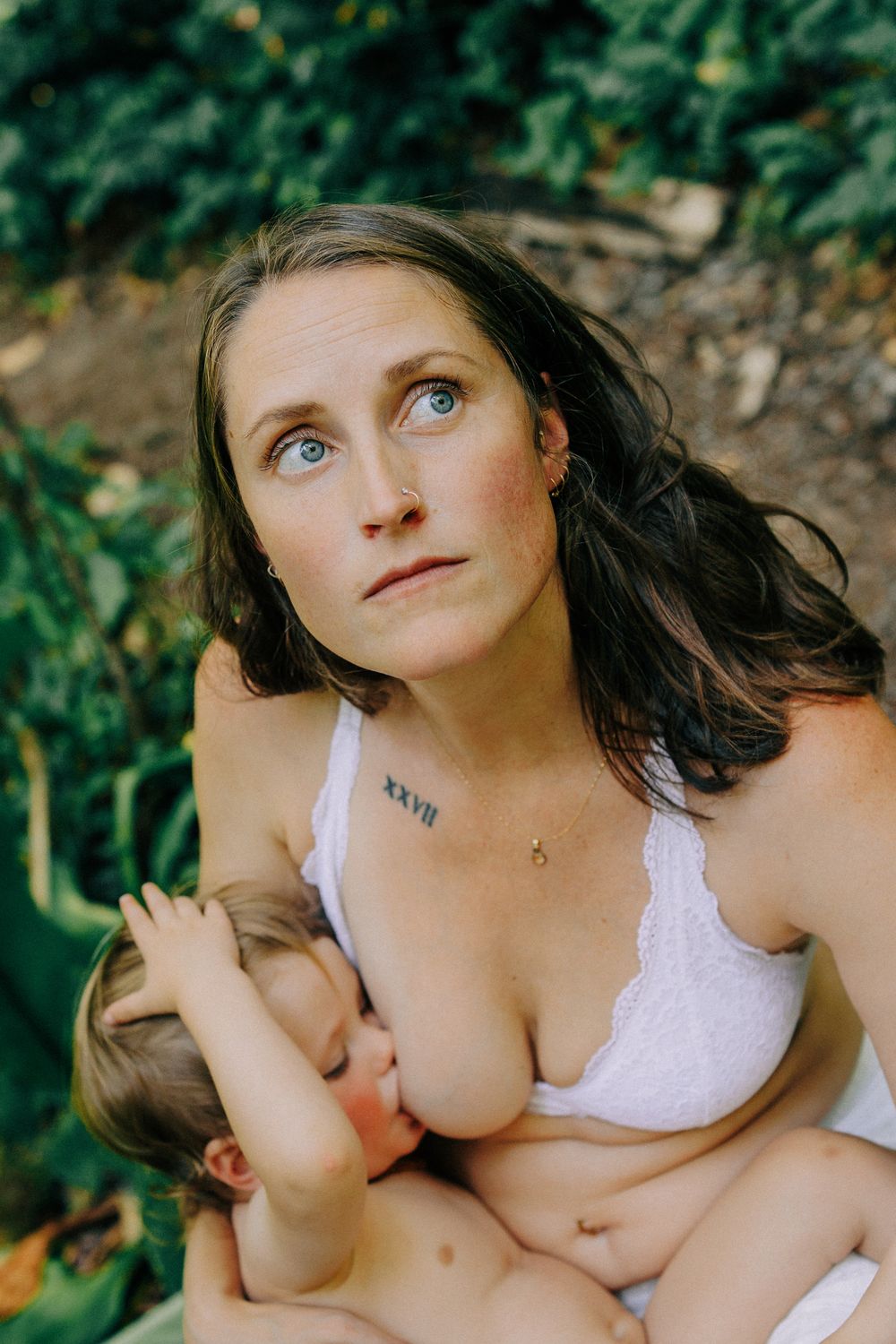 A blue-eyed mother with a tattoo breastfeeds her child outdoors in nature.