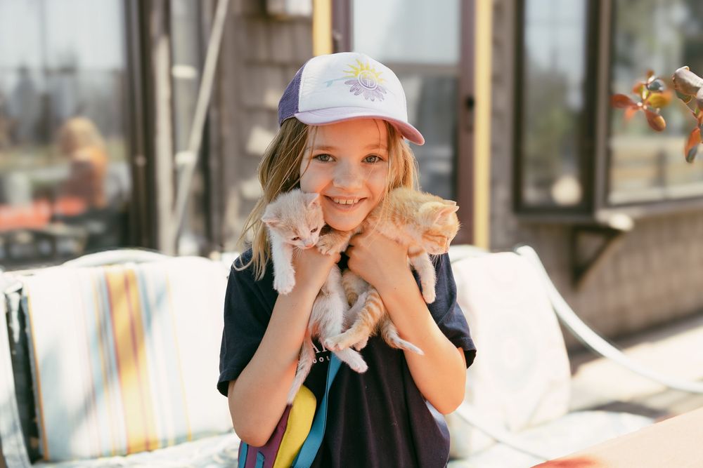 A smiling young girl holds two small kittens while wearing a baseball cap.
