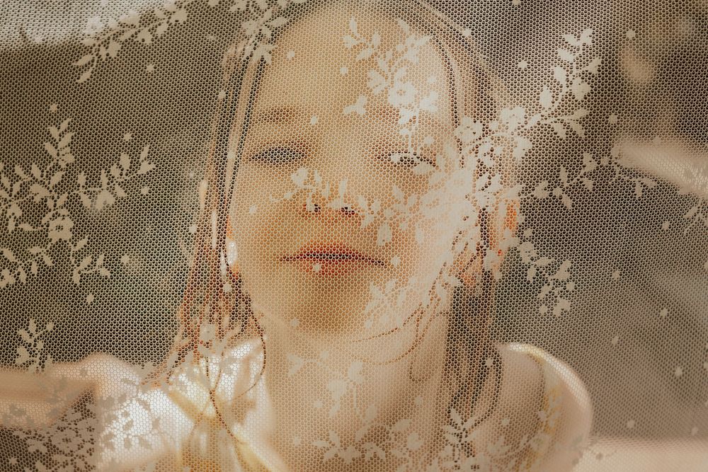 A serene young woman photographed through an embroidered floral lace fabric.