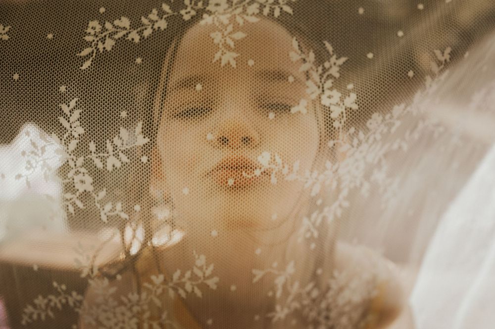 A young woman gazes upward through a delicate floral lace veil.