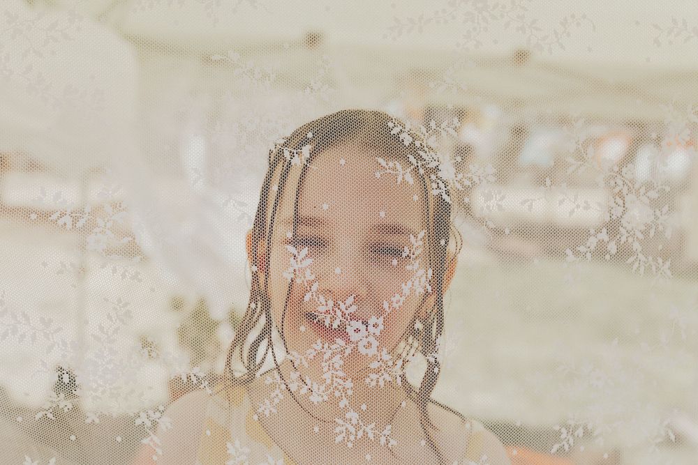 A cheerful young girl smiles through a sheer white floral embroidered fabric.