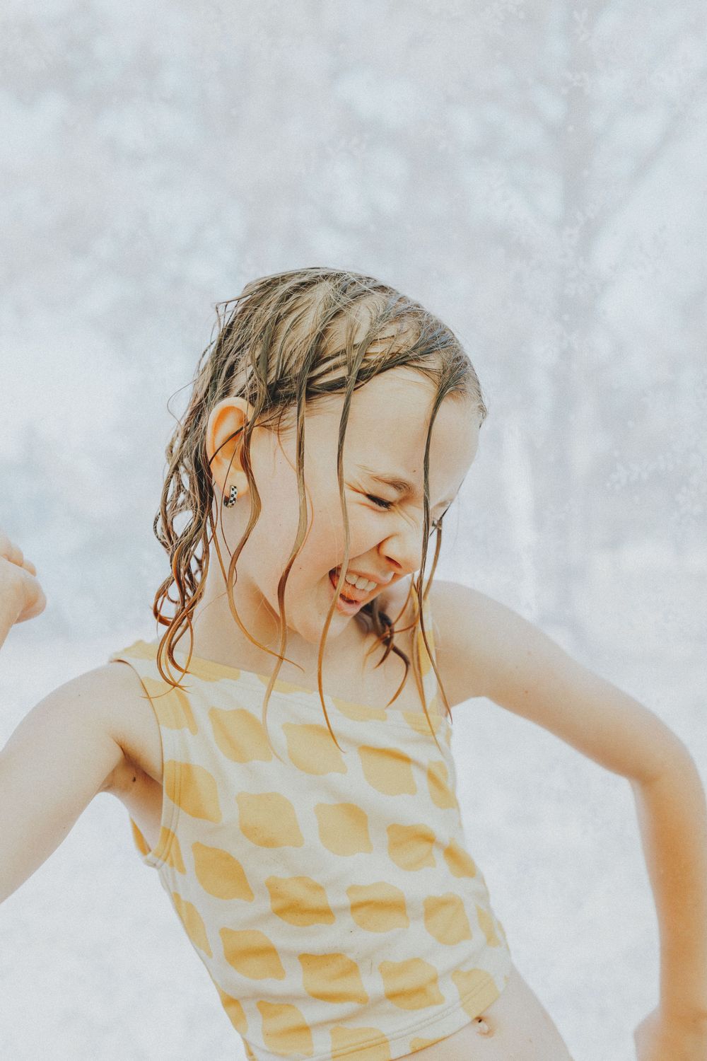 A joyful young girl with wet hair laughs while playing in water, wearing a yellow polka dot swimsuit.